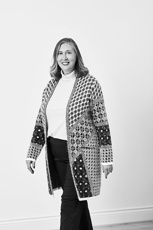 linda stands against a white background. She is smiling at the camera and wearing a long cardigan